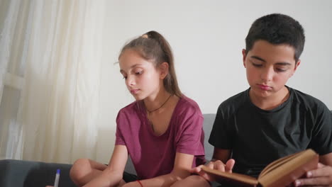 young boy seated on couch studying focused reading book while cousin beside him sits crosslegged writing attentively with pen on paper in calm indoor space filled with concentration