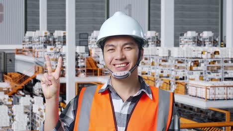 close up of asian male engineer with safety helmet standing in the warehouse with shelves full of delivery goods. smiling and showing peace gesture in the storage