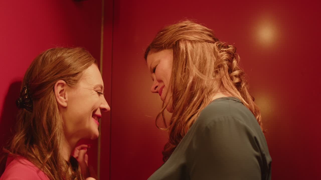 Close pan to a woman seductively whispering in the ear of another woman. They smile and walk away