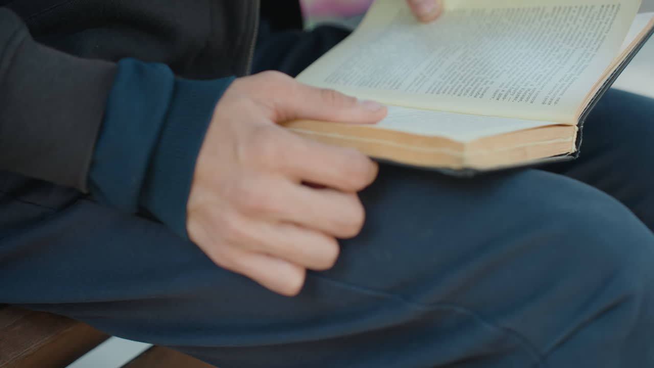 tangan membalik halaman di bangku, pria dengan celana panjang biru tua membaca buku saku usang dengan ibu jari yang lembut, bokeh taman yang diterangi matahari, bilah kayu, kertas bertekstur dan penjilidan terlihat, suasana introspeksi seorang guru pensiunan