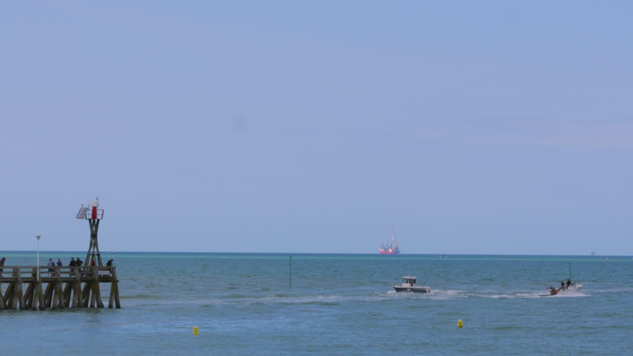 Small Boat Leaving and Entering Busy Harbour on Juno Beach with Jetty Full of Tourists and Oil Rig in Far Distance 4K