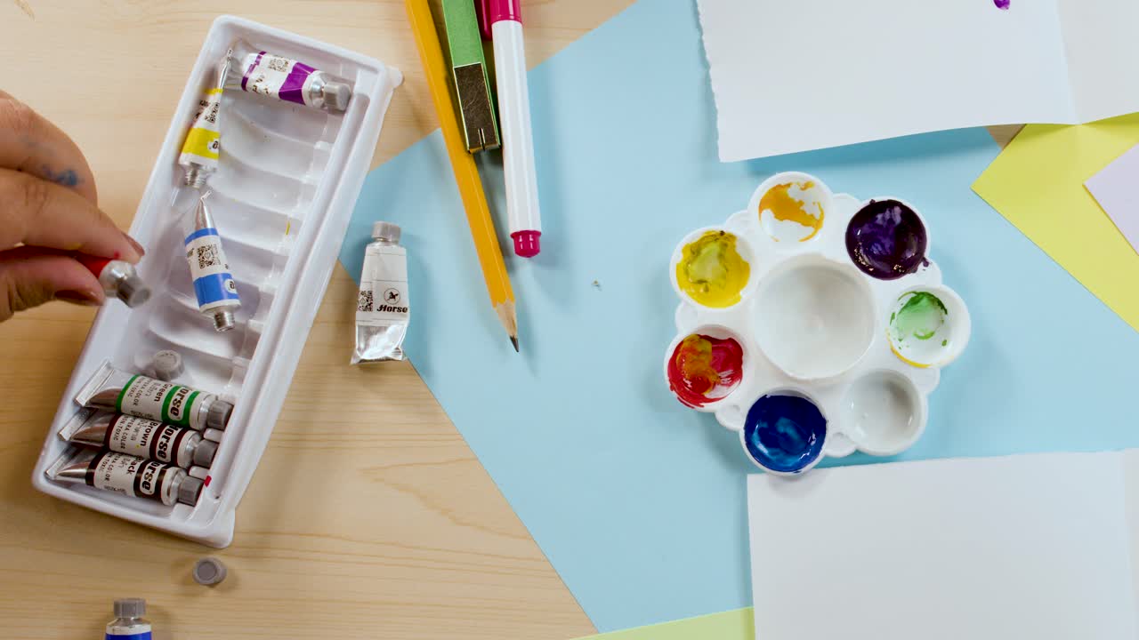 Top-down shot of hands arranging colorful paint tubes, palette, and paper on a desk
