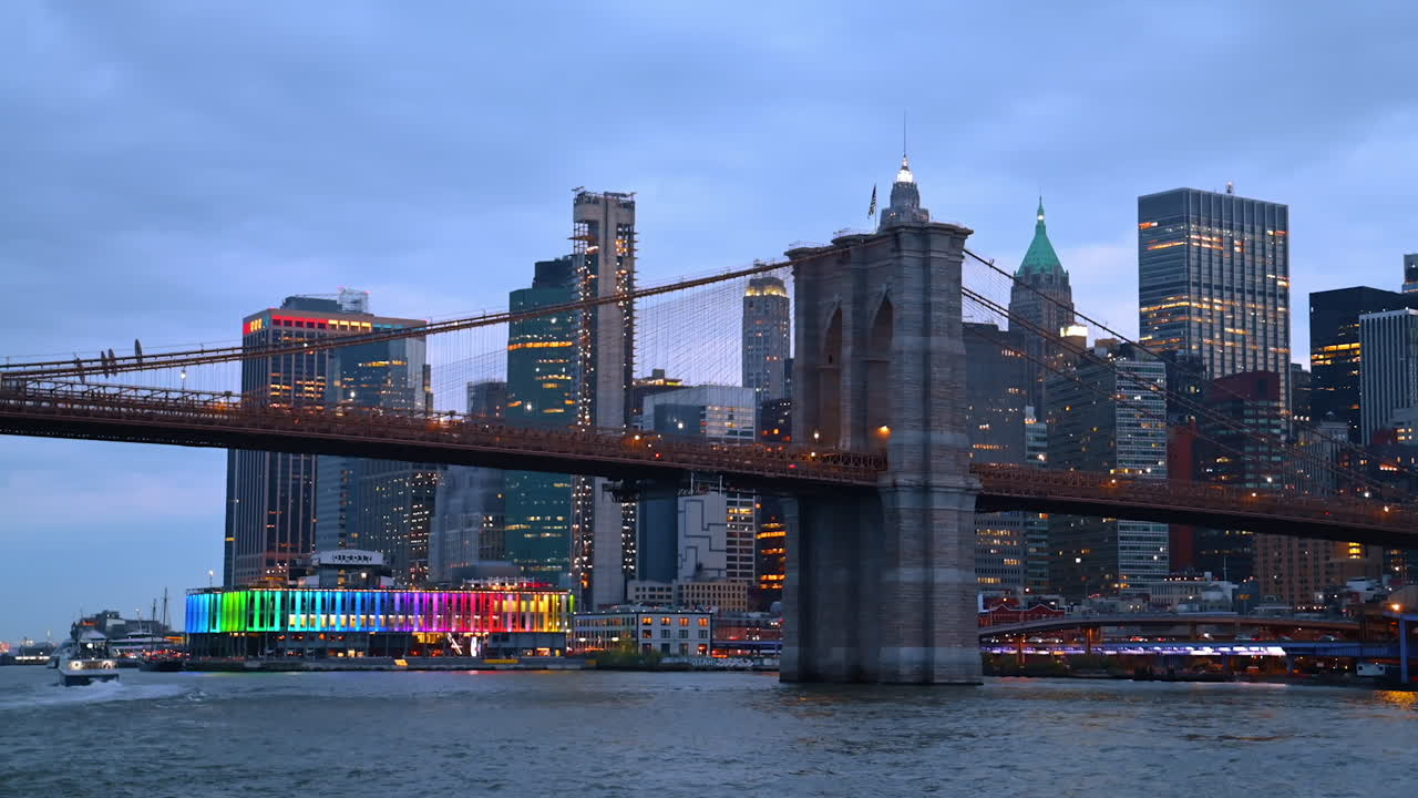 The Brooklyn Bridge at dusk time. Low angle view at the structure from the riverscape. Buildings at backdrop switching on lights