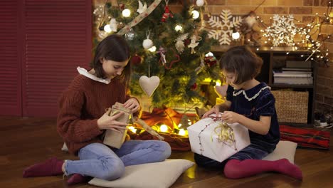 Dos-Niñas-Emocionadas-Sacudiendo-La-Caja-De-Regalo-Debajo-Del-árbol-De-Navidad-Sentadas-En-El-Suelo