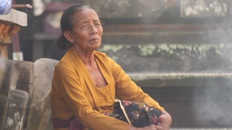 Alte-Balinesische-Frau-In-Heller-Kleidung-Sitzt-Neben-Einem-Steinaltar-Im-Mit-Rauch-Gefüllten-Innenhof-Des-Hindu-Tempels