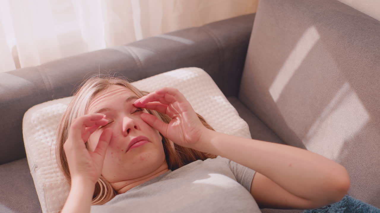Young adult lies on couch waking from sleep as she uses her hand to open her eyes and rolls them in funny playful way under soft morning sunlight filling cozy room