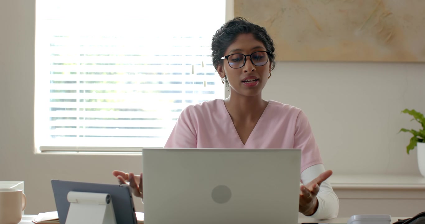 Adult African woman in pink scrub top sitting at desk reading laptop then explaining and gesturing