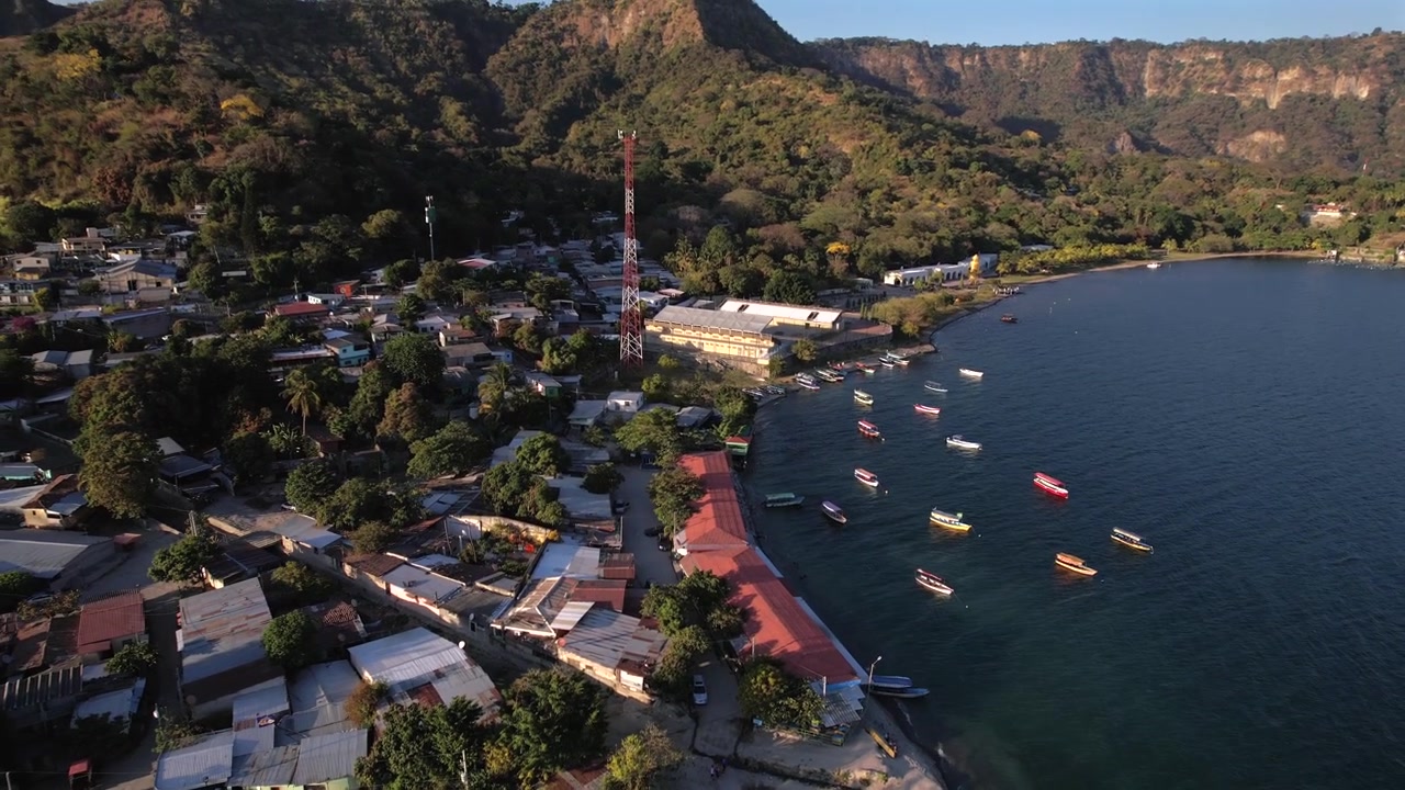 Ripresa con drone al tramonto in volo da sopra un lago verso la riva, rivelando montagne verdi, barche e case sul lago. Atmosfera tropicale e tranquilla, ideale per viaggi, turismo e narrazione naturalistica.