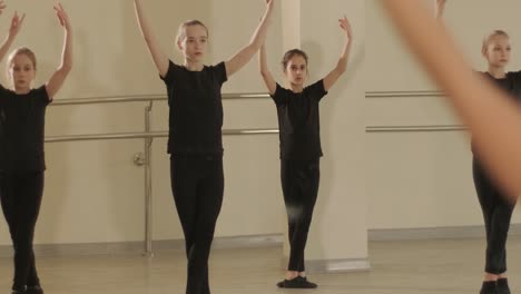 a group of young ballet students in black dancewear practicing positions in a spacious ballet studio with wooden flooring and wall-mounted barres. focused expressions and synchronized movements.