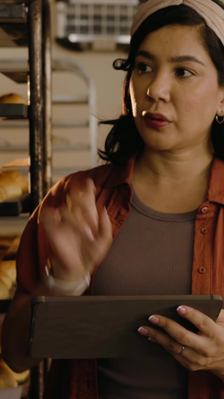 Vertical video: Pointing at trays bakery lead tapping tablet logging near rack and ovens rust top