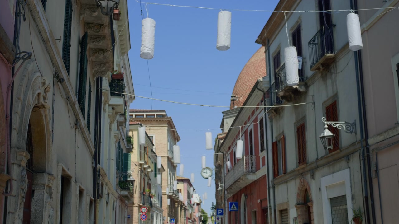 Old town street hanging lanterns, handheld panning shot looking down a charming narrow Italian street decorated with white cylindrical paper decorations under a clear blue sky during bright daylight.