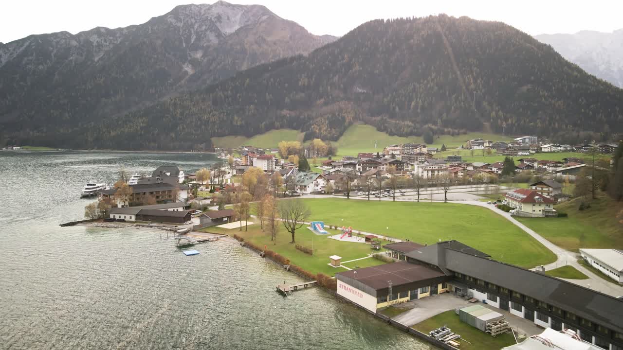A stunning drone shot from Achensee Pertisau, flying towards the picturesque lake town. The vibrant turquoise waters and majestic alpine surroundings create a breathtaking scene in Austria.