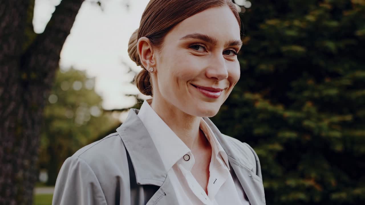 Professional businesswoman standing confidently in park setting, wearing light gray trench coat and crisp white shirt, radiating executive elegance during golden hour sunlight