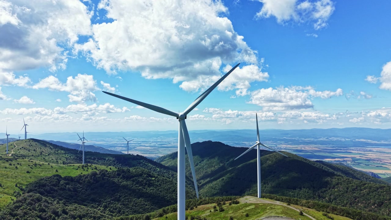 Eoliche su una collina sotto un cielo azzurro con nuvole in un paesaggio con campi verdi e montagne distanti durante una giornata di sole.