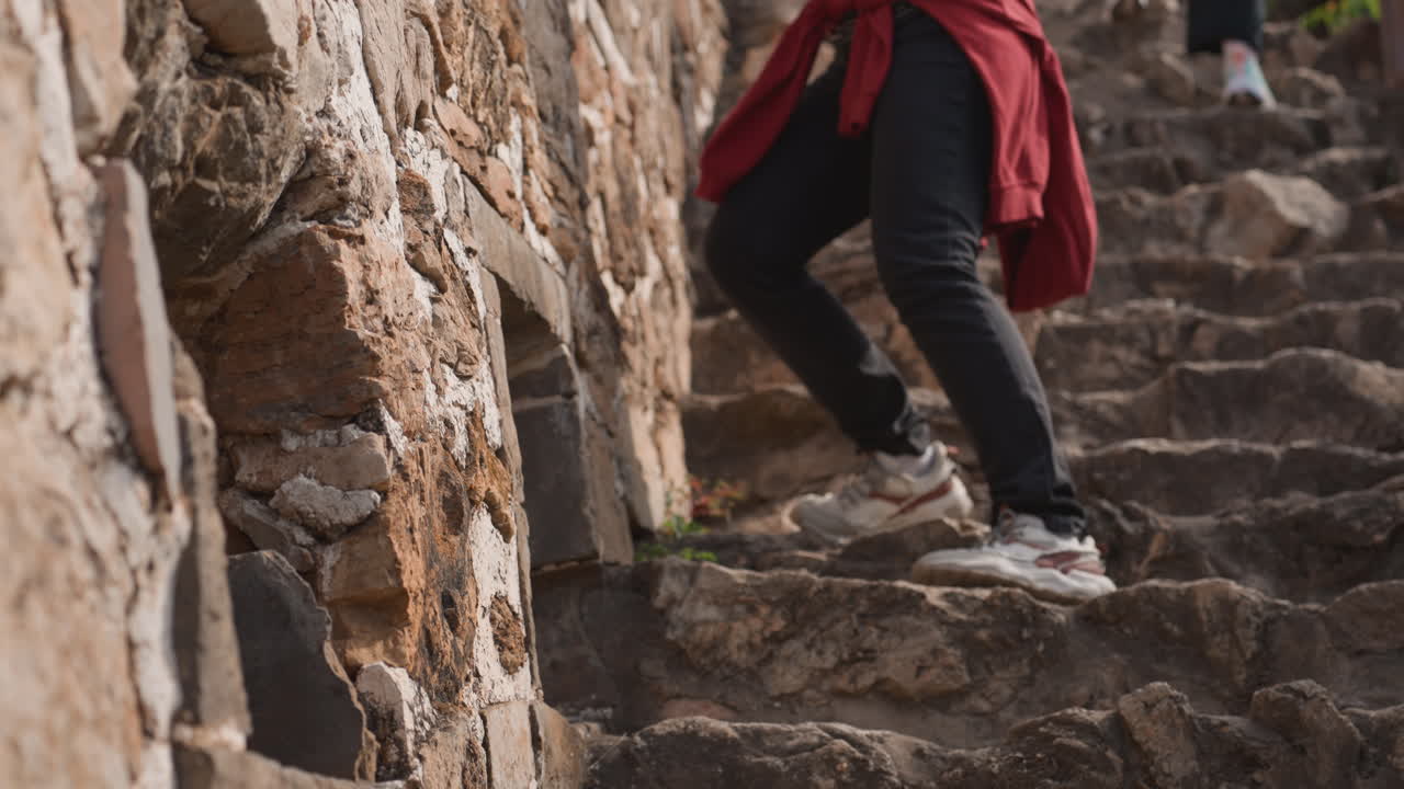 Woman climbing stone stairs at ruins. Closeup of hands bracing against rough masonry while female traveler with red jacket tied at waist and backpack navigates uneven steps companion follows