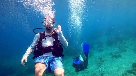 scuba-diver-having-fun,-making-a-joke,-doing-the-OK-sign-or-OK-gesture,-scuba-diving-in-swimshorts,-sunny-tropical-ocean-water,-underwater-seaworld,-cinematic-colorful-medium-wide-shot