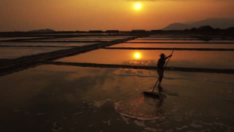 worker doing overtime in the golden evening light silhouetted racking the salt field for harvest in slow motion