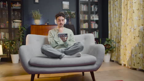 young man watching comic movie on smartphone while sitting with crossed legs on couch at home