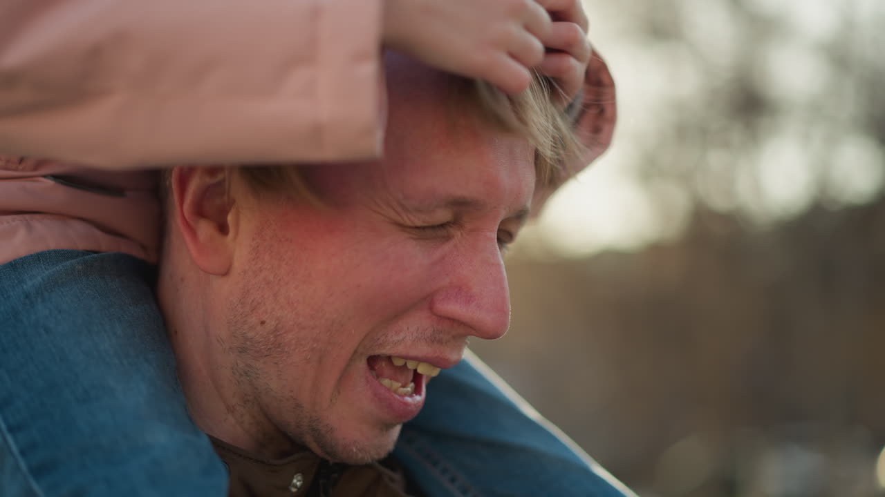 primer plano de un padre alegre mientras su hija pequeña, sentada en sus hombros, toca juguetonamente su cabello en un parque