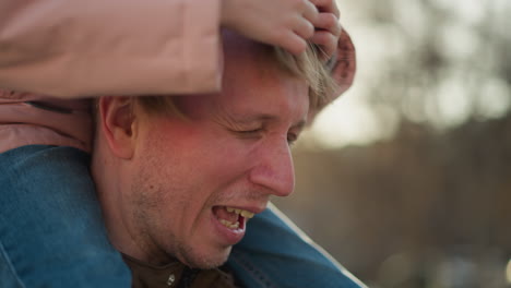 close-up of a joyful father as his little daughter, sitting on his shoulders, playfully touches his hair in a park setting