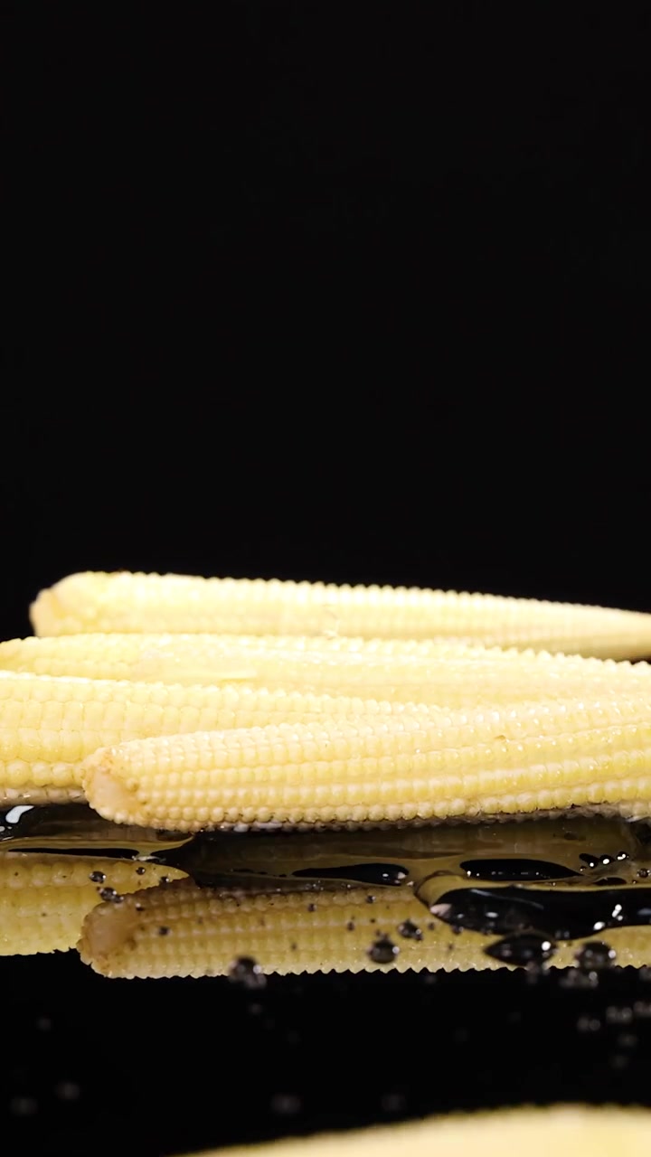 Several baby corn cobs fall and land on a glossy, wet black surface, creating water splashes. Studio lighting highlights texture and reflections