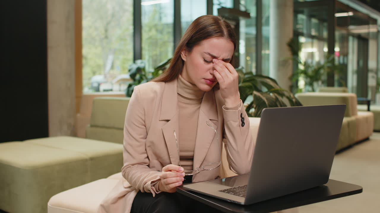 Overworked businesswoman suffering from eye strain and headache working on laptop in office lobby