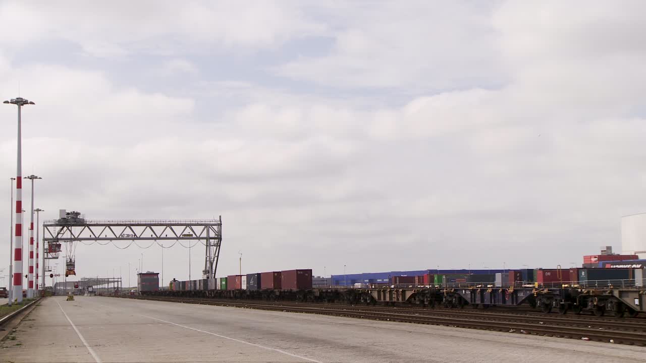 From a parallel road perspective, an overcast train yard manifests, strewn with multicolored cargo containers alongside tracks, with a towering crane in backdrop