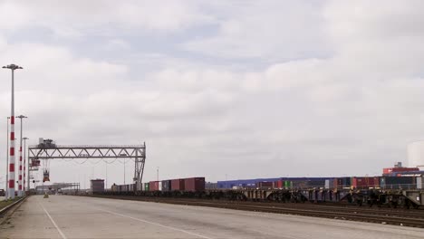 from a parallel road perspective, an overcast train yard manifests, strewn with multicolored cargo containers alongside tracks, with a towering crane in backdrop