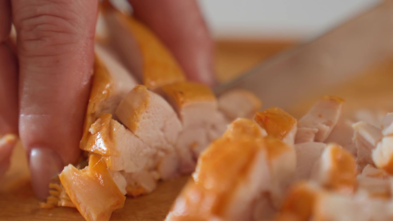 Primo piano di preparazione in cucina che mostra mani che affettano pollo cotto in pezzi sottili e lucidi su un tagliere di legno, con un contatto attento della lama, movimento guidato, luce interna calda e ricchi dettagli di texture.