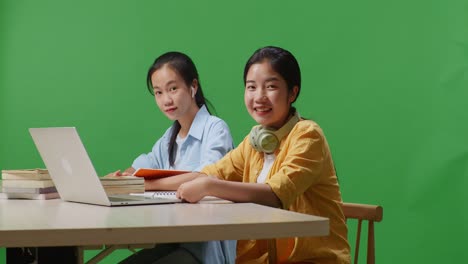 asian woman students reading books and showing thumbs up gesture to the camera while sitting on a table with laptop in the green screen background classroom