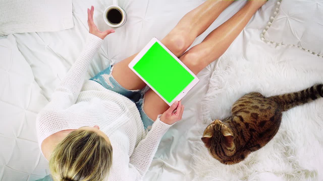 vista de arriba hacia abajo de una mujer sentada en la cama usando un dispositivo de tableta con una pantalla verde en blanco