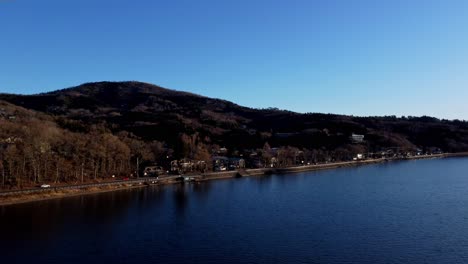 A-serene-lakeside-with-houses,-trees,-and-a-hill-during-early-evening,-aerial-view