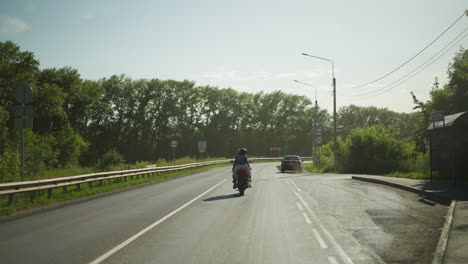 back view of two friends riding a power bike slowly down the road, with a car ahead emitting smoke from its exhaust, trees and street signs are visible in the background