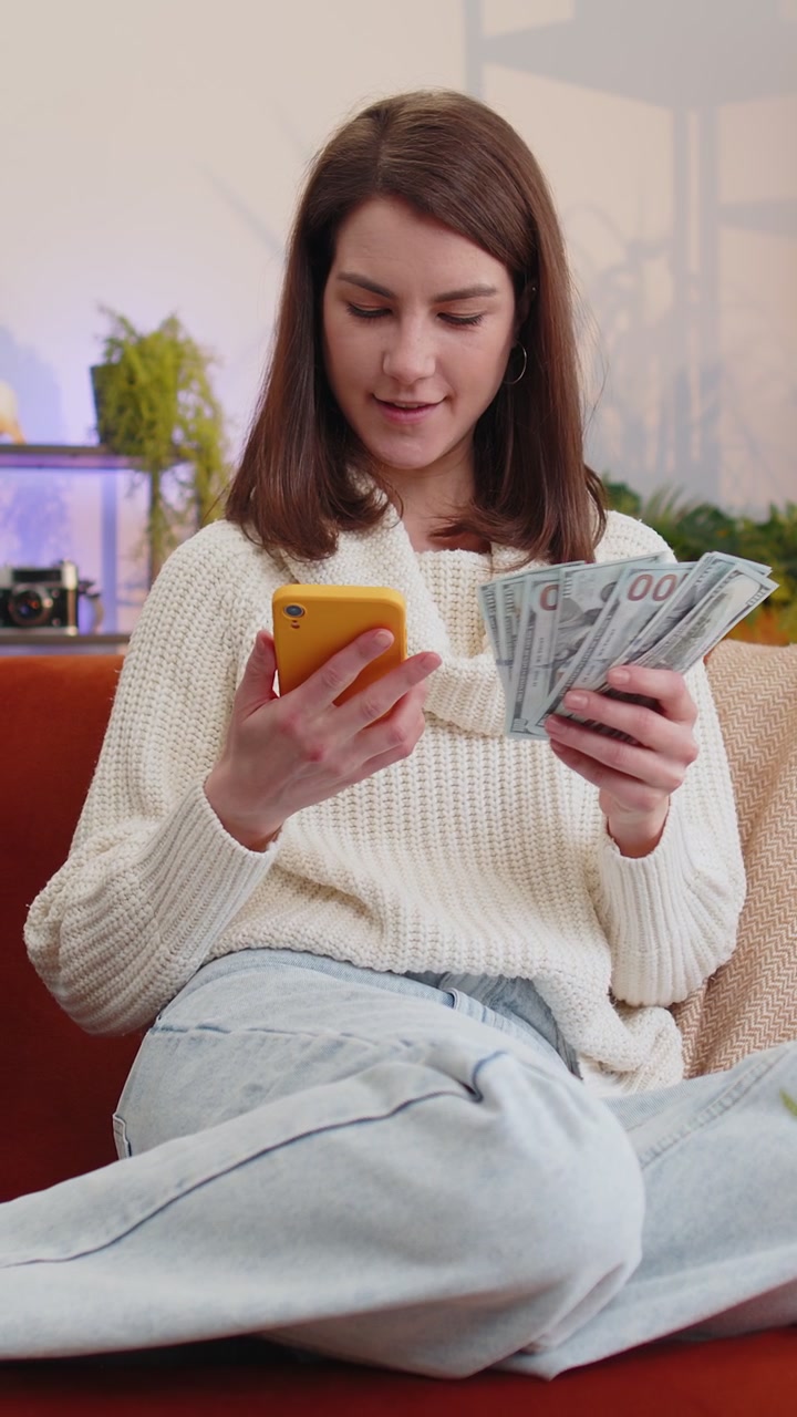 Una giovane donna sorridente e felice che conta i soldi in contanti usa lo smartphone il reddito risparmia il budget della lotteria a casa