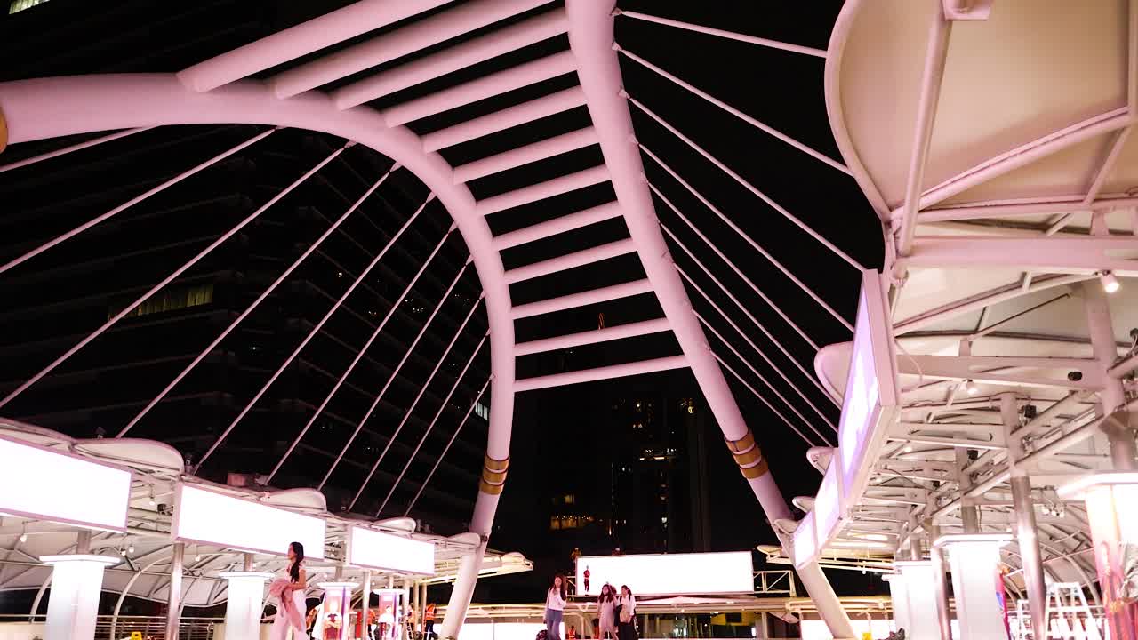 People walk beneath illuminated Silom Bridge at night, vibrant city lights reflecting on wet pavement