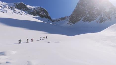 Aerial-revealing-shot-showing-group-of-mountaineer-hiking-uphill-snowy-mountains-in-sunlight