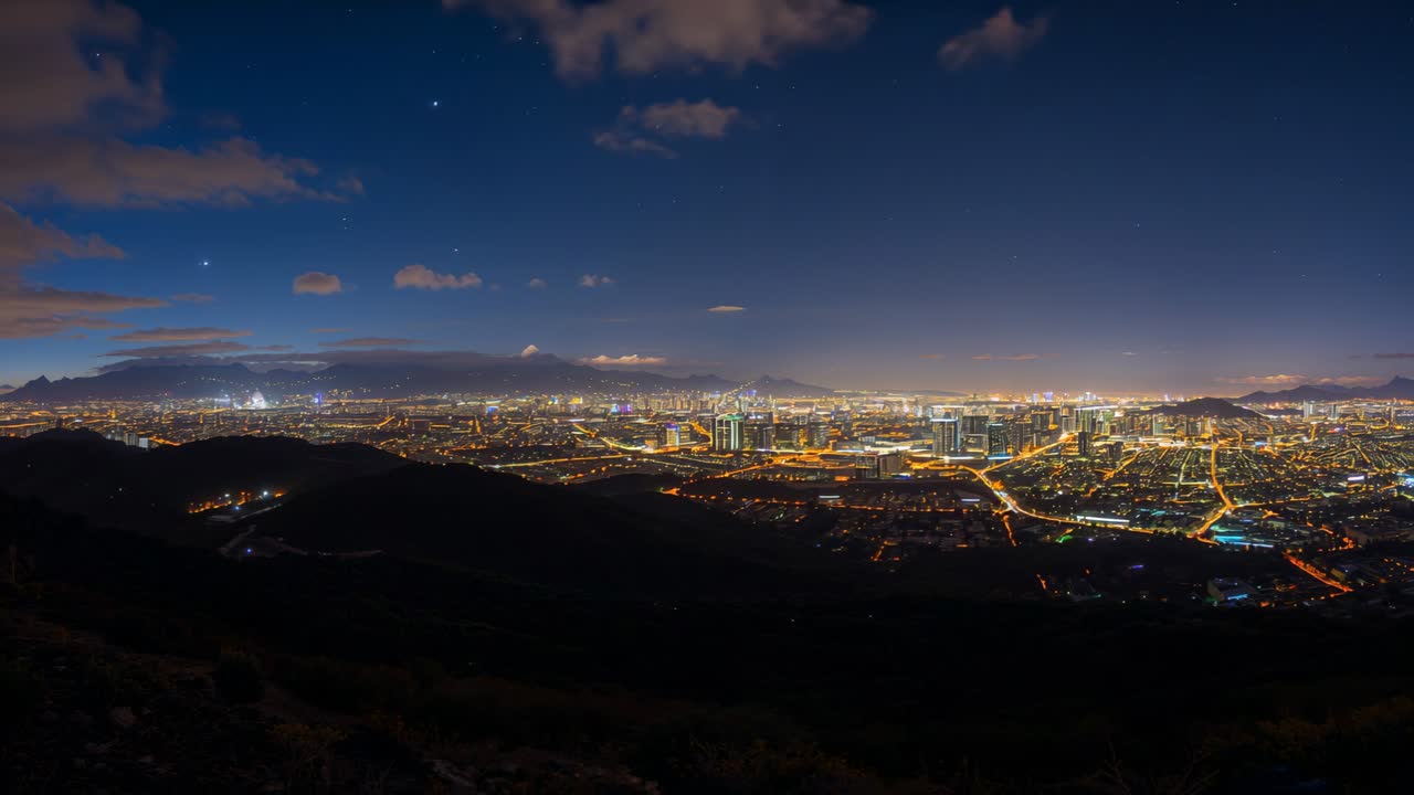 El perfil urbano de la ciudad brilla, con carreteras iluminadas proyectando un resplandor en el horizonte desde una colina, mientras las nubes a la deriva iluminan el cielo.