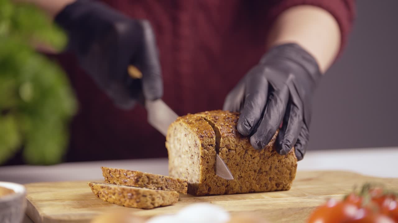 Slowmotion closeup of a chef slicing whole grain bread on a wooden board. The sharp knife glides smoothly, revealing a crisp crust Black gloves ensure hygiene, with fresh ingredients in the background