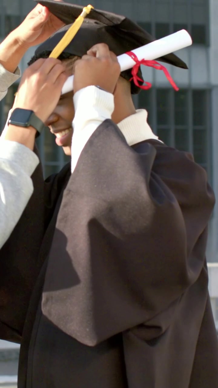Vertical video: Reaching hooded pal adjusting grad gown, cap on roof moving yellow tassel for grad