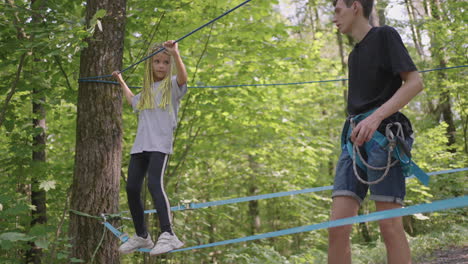 small girl in climbing equipment in a rope park. group of caucasian children training at boot camp. in the children camp children are taught to overcome obstacles with the help of a rope crossing