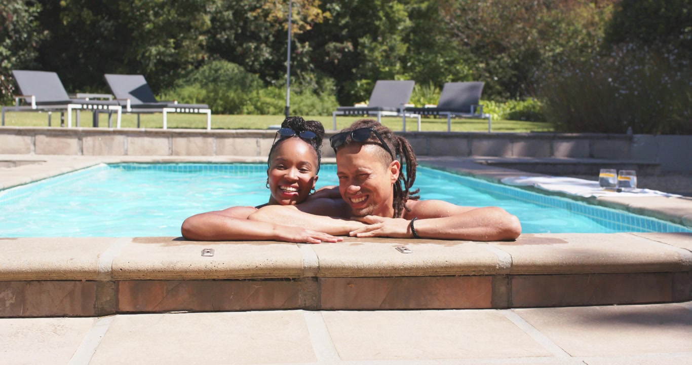 Pareja africana apoyada junto a la piscina, mujer en traje de baño ajustándose las gafas de sol, hombre sin camiseta tocando, sonriendo.