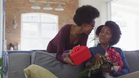 Happy-african-american-mother-and-daughter-sitting-on-sofa,-giving-roses-and-present