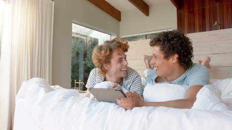 una pareja gay feliz y diversa acostada en la cama y usando una tableta en casa, en cámara lenta