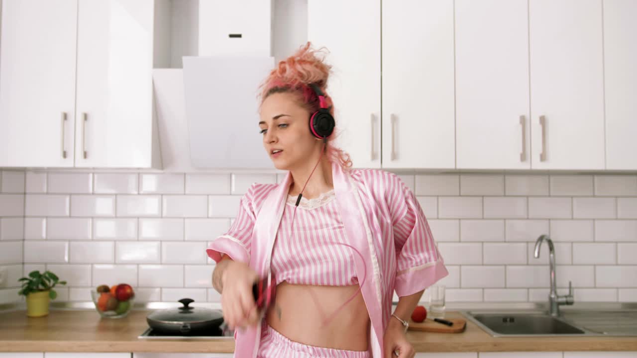 joven feliz con cabello rosa bailando en la cocina con pijama rosa y escuchando música con auriculares