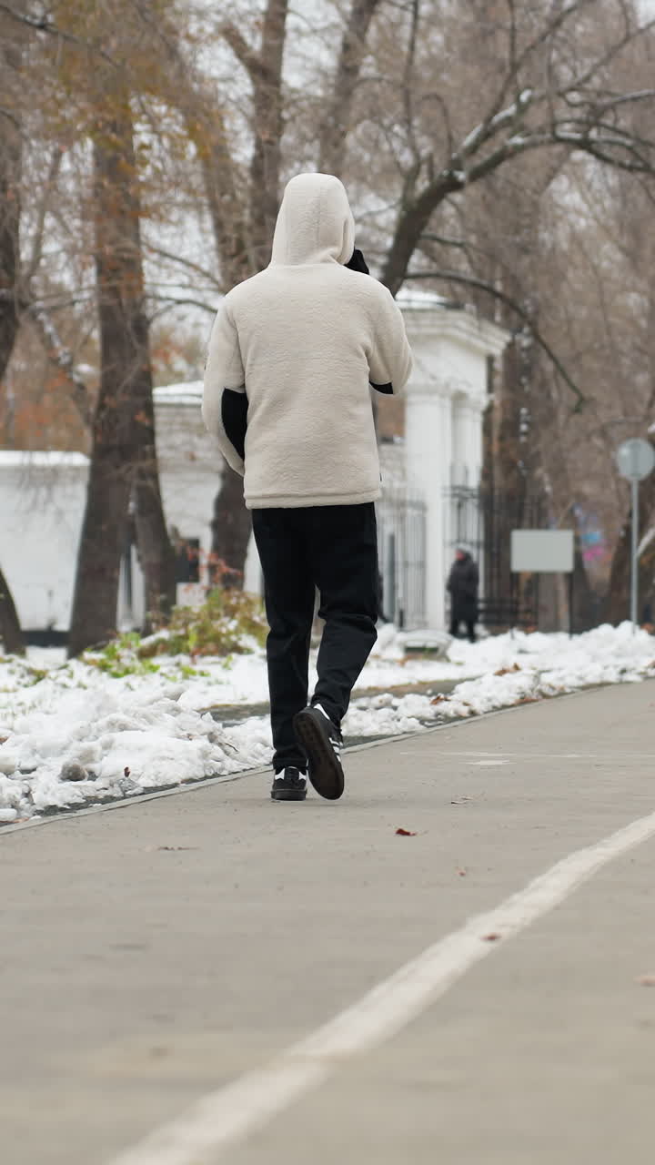 전화 통화 중인 젊은 남자의 뒷면은 겨울에 거리를 걸어다니며 자전거를 타고 다가오는 누군가의 흐릿한 모습, 바닥의 눈과 배경의 나무입니다.