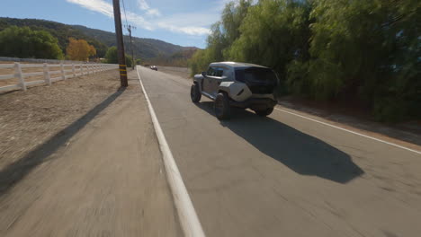 fpv drone cobertura cinematográfica de 180 grados del tanque rezvani de grado militar desde los lados, siguiendo una carretera rural cubierta de árboles cerca de un pequeño pueblo