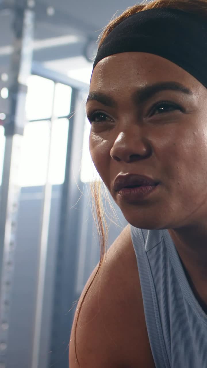 Vertical video: Panting after set, woman wearing black headband and tank top coaching at gym rack