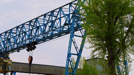 metal blue crane with automated hook in the middle and hanging cable. green tree growing at the territory beside the crane.