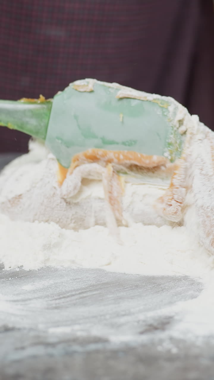 Primo piano di un fornaio che mescola un impasto denso con la farina su un piano di lavoro usando una spatola verde; mani che impastano e piegano il composto per preparare dolci fatti in casa