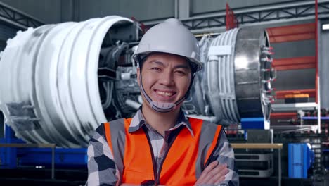 close up of asian male engineer with safety helmet crossing his arms and smiling to camera while standing with airplane engine maintenance conducted, analytics checking the turbine